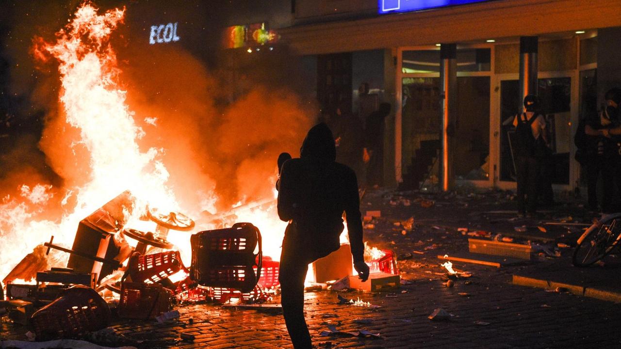 Vor dem Supermarkt mit zerborstenen Scheiben brennt im Dunkeln eine Barrikade; ein Demonstrant kickt etwas in das Feuer.