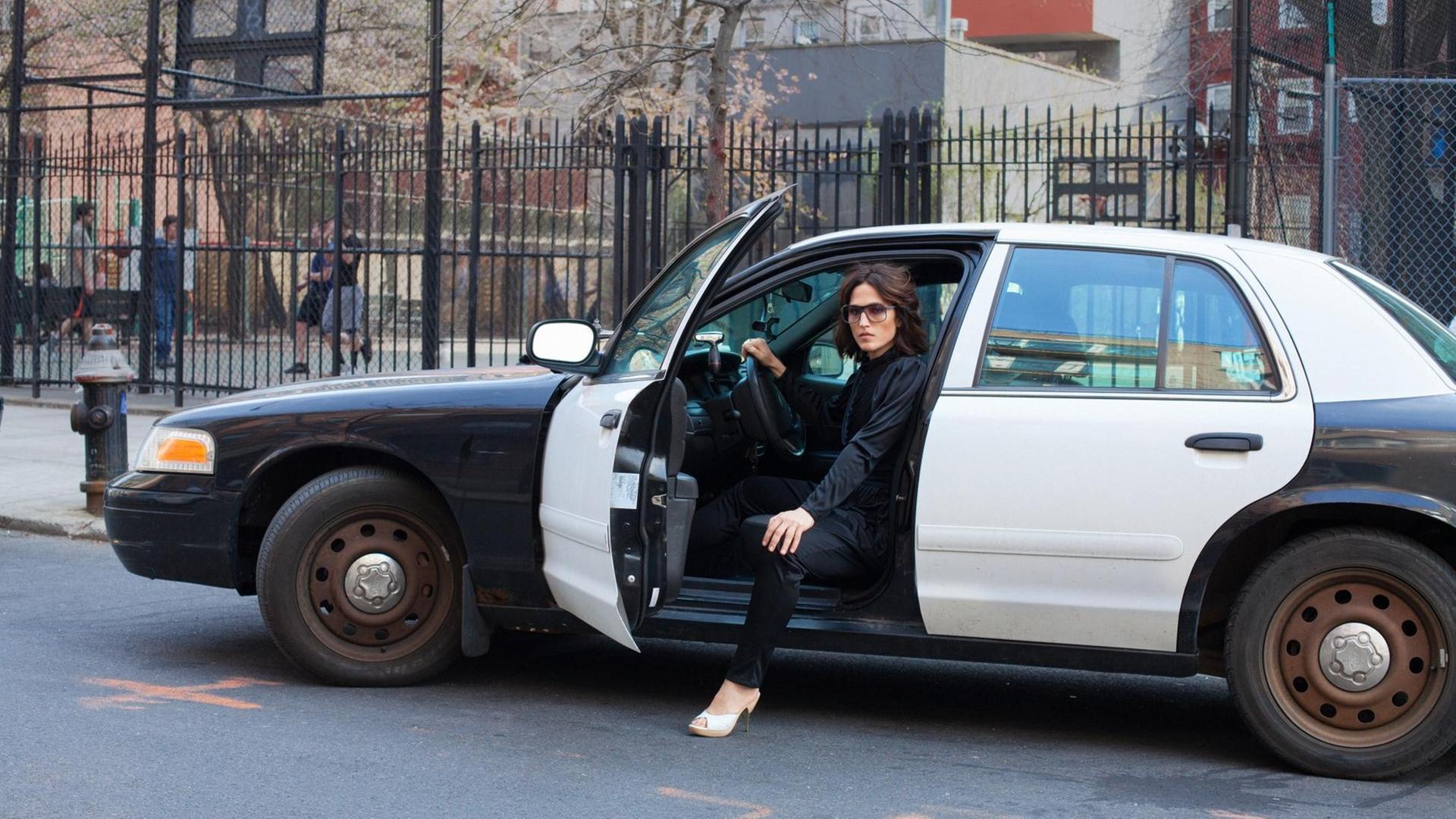 Eine Frau sitz am Steuer eine alten amerikanischen Polizeiautos. Die Autotür ist geöffnet. Ein Bein stellt sie auf dem auf dem Boden.