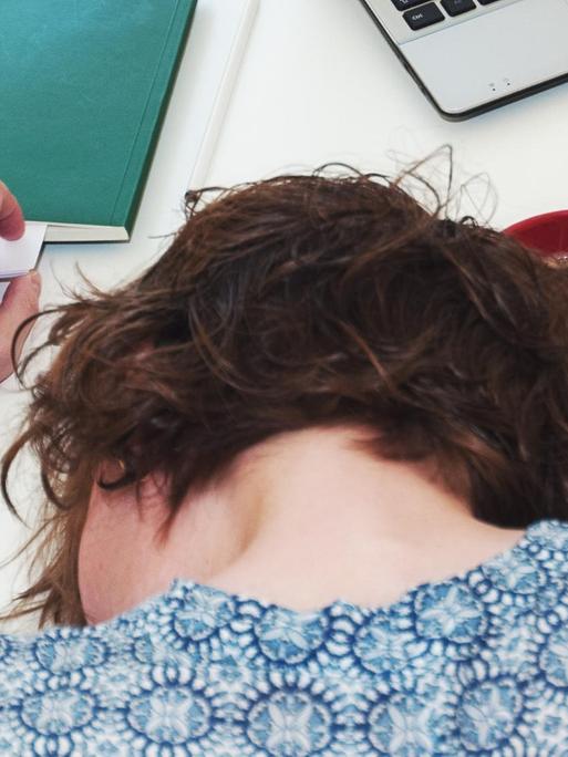 Eine Frau liegt mit dem Kopf auf einem Tisch neben ihrer vollen Kaffeetasse und ihrem Rechner.