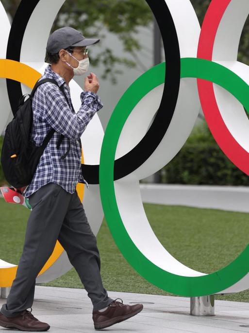 Ein Mann mit Maske läuft an einer Installation Olympischer Ringe vorbei, die in Tokio an die wegen Corona verschobene Sportveranstaltung erinnert.