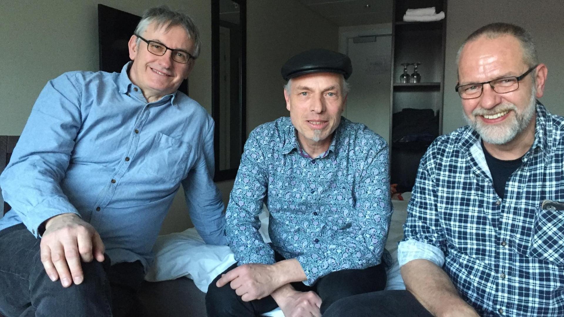 Drei Männer sitzen auf einem Hotelbett und blicken in die Kamera