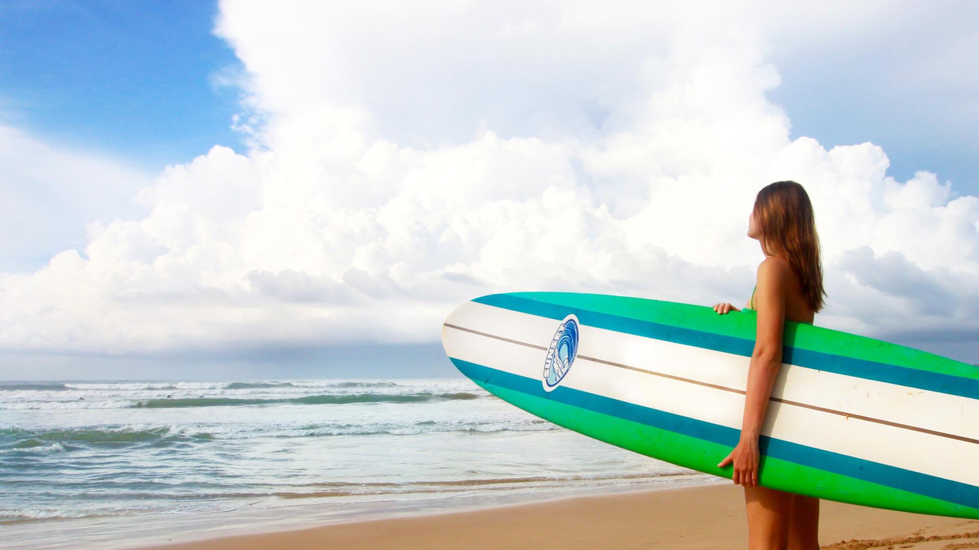 Eine Frau steht mit einem Surfbrett unter dem Arm am Strand und guckt auf das Meer hinaus.