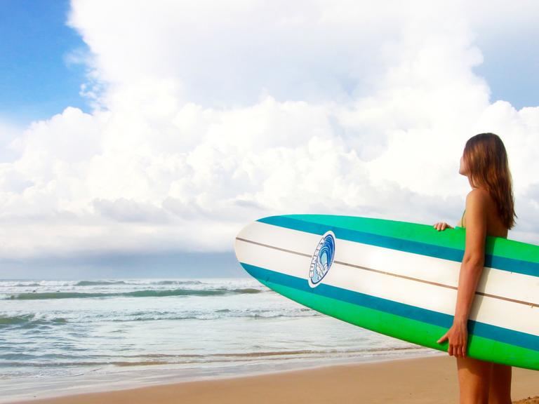 Eine Frau steht mit einem Surfbrett unter dem Arm am Strand und guckt auf das Meer hinaus.