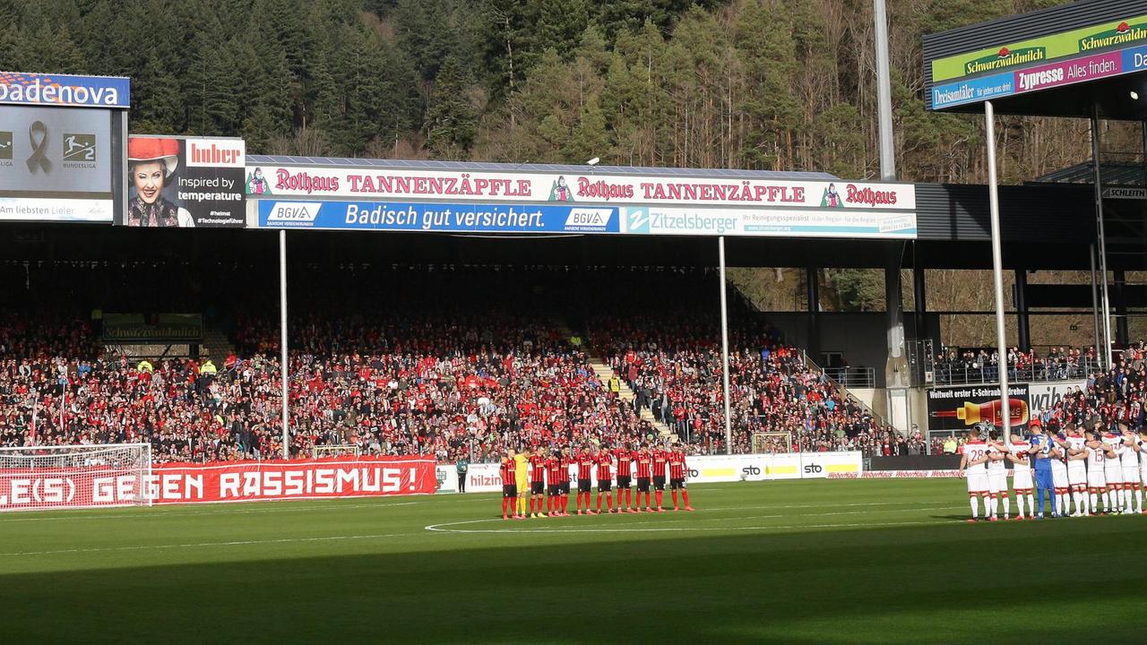 Zu sehen ist das Freiburger-Fußball-Stadion. Auf dem Rasen stehen in einem Halbkreis die Spieler von Freiburg und Düsseldorf, auf der Leinwand wird den Opfern des Anschlags in Hanau gedacht.