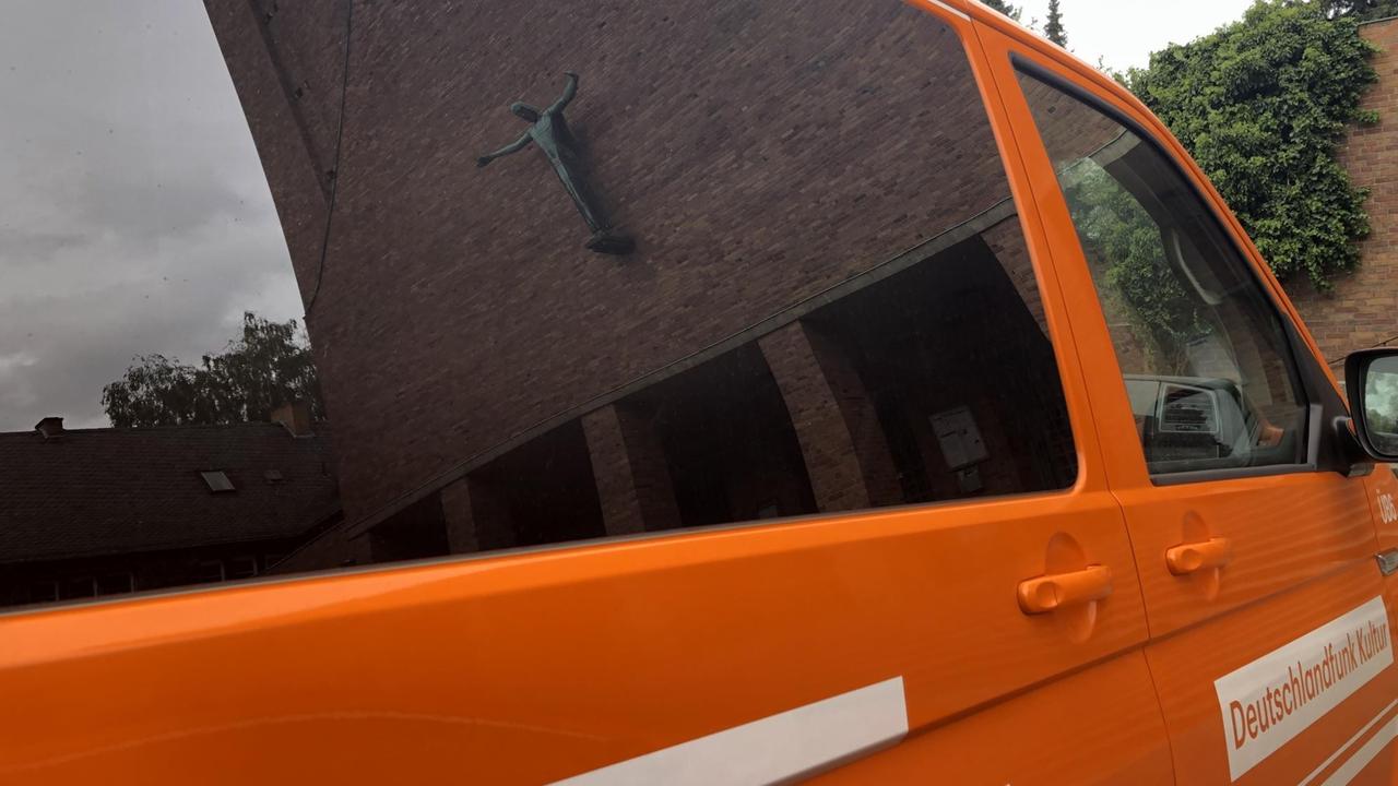 In den Autoscheiben unseres orange-farbenen Kleinbusses spiegelt sich die bronzene Jesusfigur der Außenwand der Kirche.