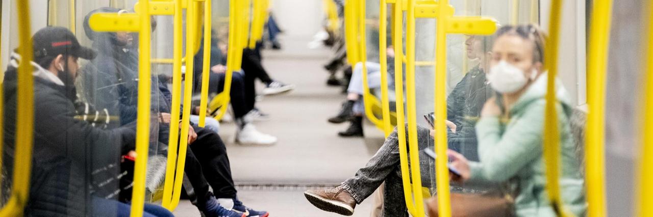 Fahrgäste sitzen in einer U-Bahn, wobei eine Frau einen Mundschutz trägt. 