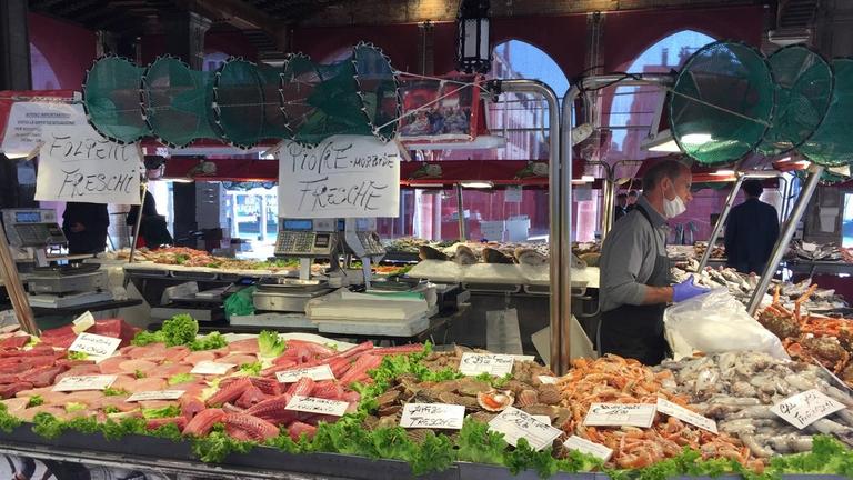 An einem Marktstand liegen viele verschiedene Fischsorten in der Auslage. Ein Verkäufer mit Schürze unterhät sich mit seiner Kundschaft.