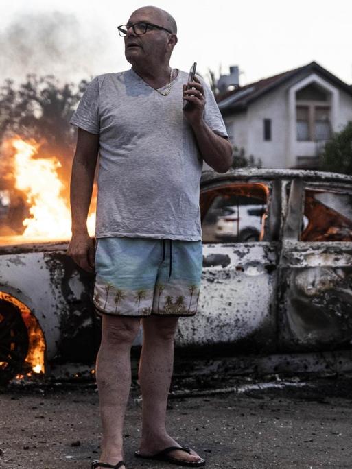 Der Mann in Flipflops, Badehose und T-Schirt steht vor dem brennenden Wrack auf der Straße und hält ein Handy.