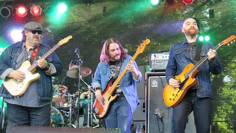 Zwei Gitarristen, ein Bassist und ein Trommler stehen auf einer Bühne und spielen Bluesmusik.