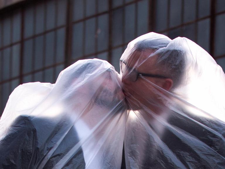 Die beiden Musiker, jeweils mit einer Plastiktüte über dem Kopf, küssen sich.