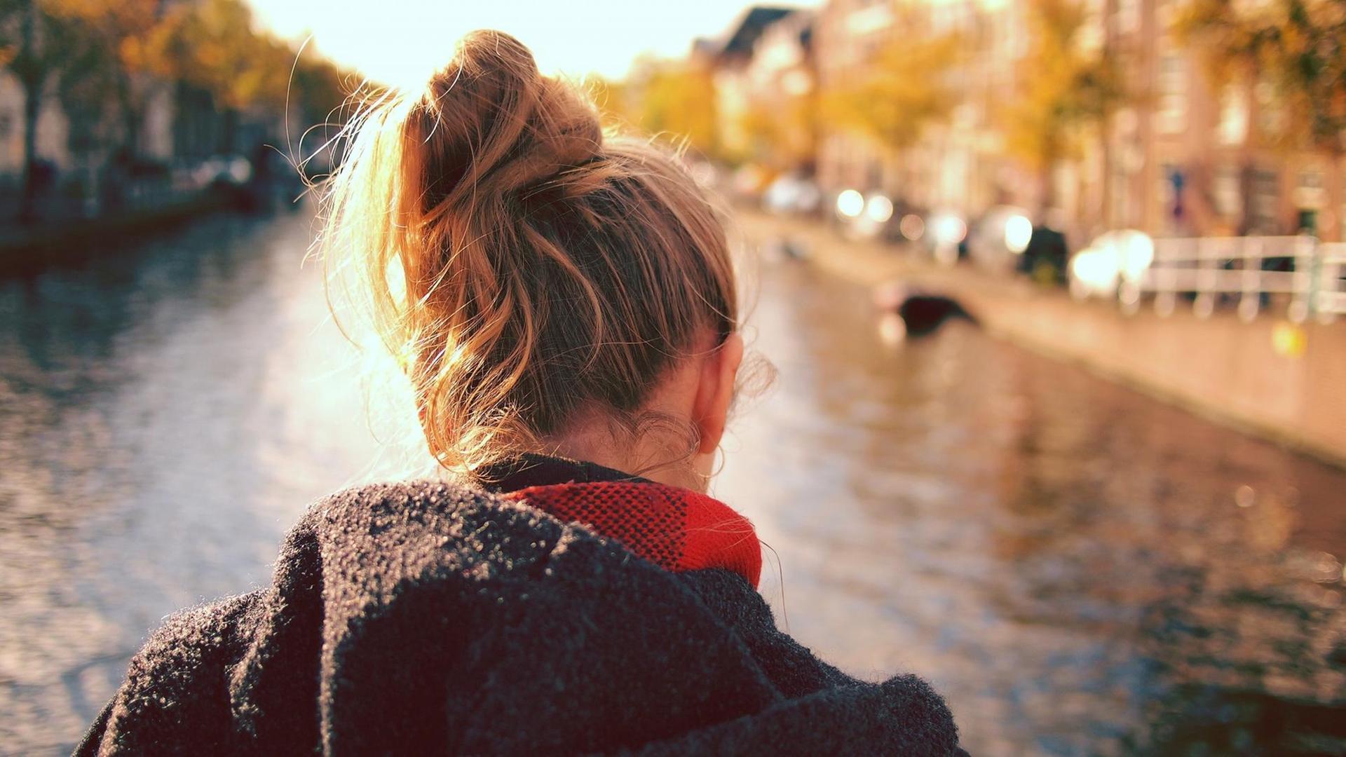 Das Foto zeigt eine blonde junge Frau mit Dutt von hinten. Sie blickt auf einen Fluss. Rechts und links ist das Flussufer zu sehen.