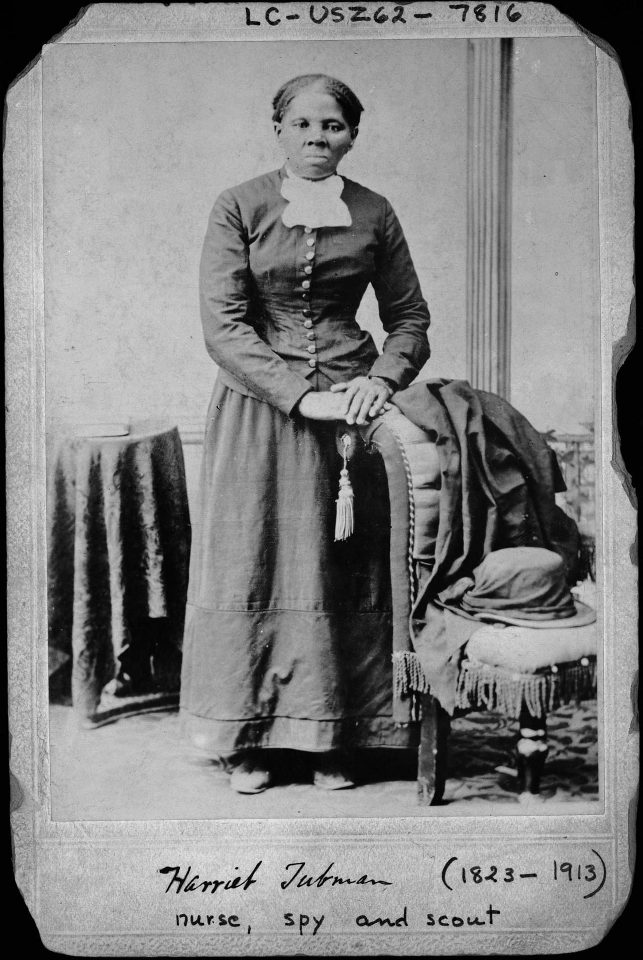 Harriet Tubman, stehend, grimmig schauend, in einem hochgeschlossenen Kleid und auf einen Stuhl gestützt.