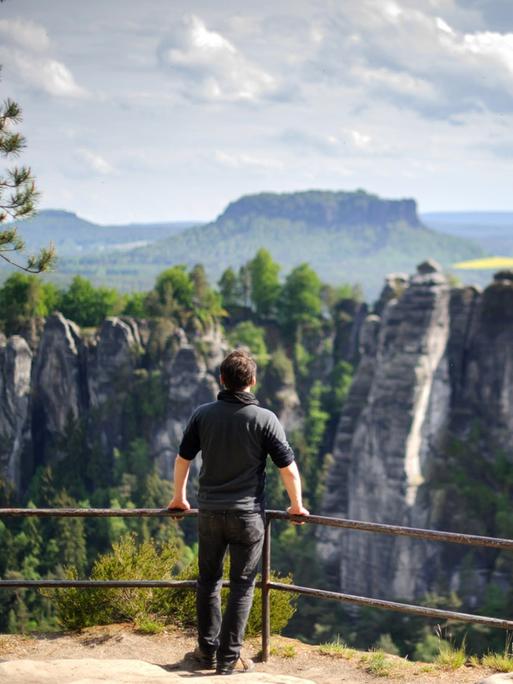 Ein Wanderer steht am 19.05.2015 in der Nähe von Rathen (Sachsen) auf einem Felsen in der Nationalparkregion Sächsische Schweiz und schaut zur Bastei hinüber, im Hintergrund erhebt sich der Lilienstein.