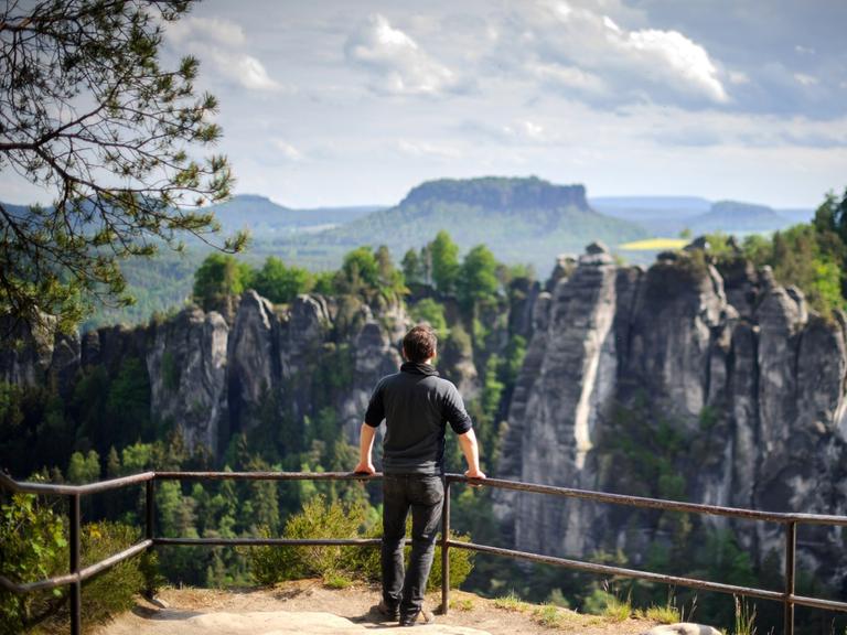 Ein Wanderer steht am 19.05.2015 in der Nähe von Rathen (Sachsen) auf einem Felsen in der Nationalparkregion Sächsische Schweiz und schaut zur Bastei hinüber, im Hintergrund erhebt sich der Lilienstein.