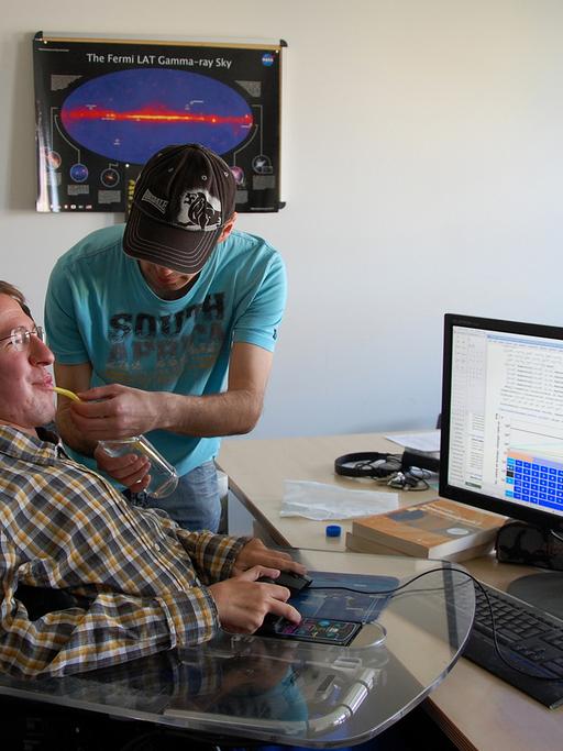 Der körperlich stark behinderte Christoph Wendel sitzt in einem Rollstuhl vor einem Computer, während sein Assistent Alex ihm eine Flasche mit Strohhalm zum Trinken an den Mund hält.