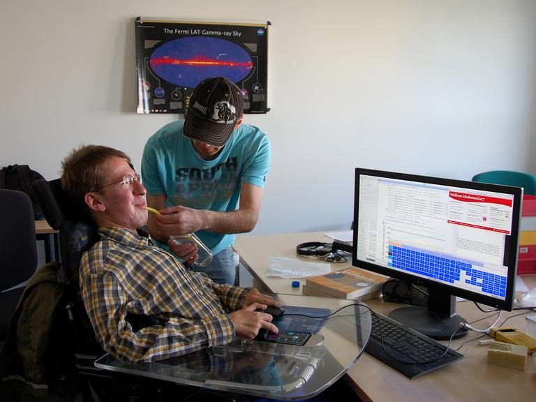 Der körperlich stark behinderte Christoph Wendel sitzt in einem Rollstuhl vor einem Computer, während sein Assistent Alex ihm eine Flasche mit Strohhalm zum Trinken an den Mund hält.