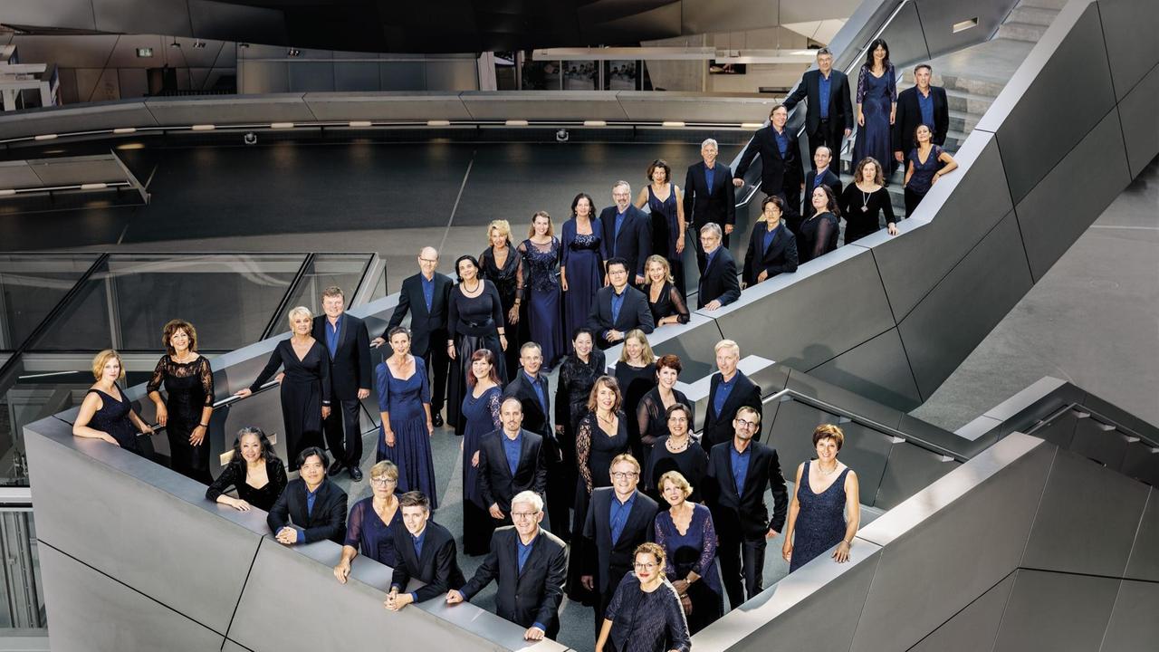 Der Chor steht in Konzertkleidung in einem modernen Treppenhaus, das auf grauen, kantigen Linien beruht.
