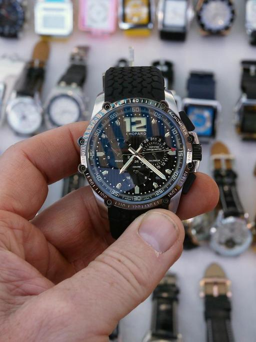Eine Hand hält eine plagiierte Uhr der Marke Chopard in die Kamera, darunter sind in einer Auslage zahlreiche weiterer nachgemachte Uhren zu sehen