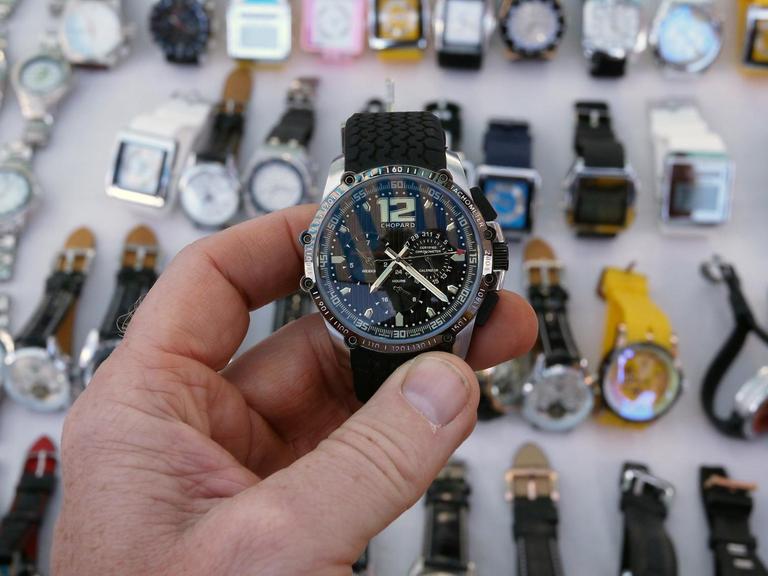 Eine Hand hält eine plagiierte Uhr der Marke Chopard in die Kamera, darunter sind in einer Auslage zahlreiche weiterer nachgemachte Uhren zu sehen