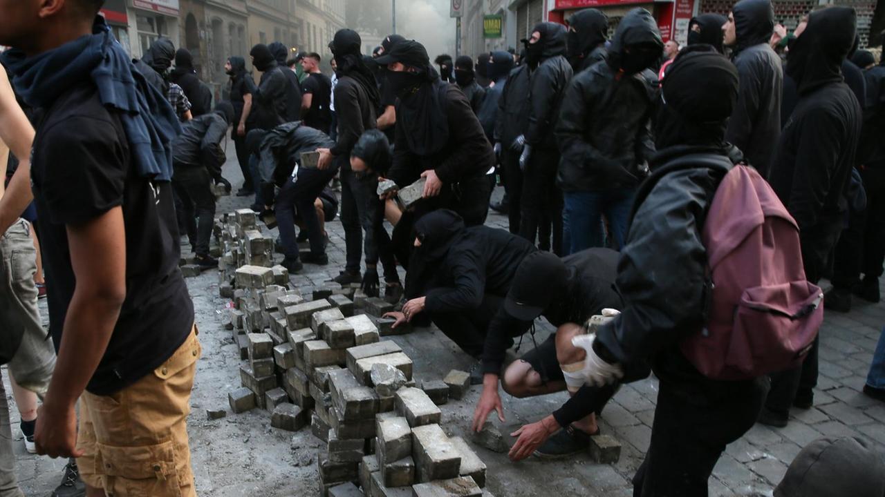 Die schwarzgekleideten und teils vermummten Demonstranten errichten mehrere Stapel Pflastersteine, die sie aus dem Straßenbelag ziehen.
