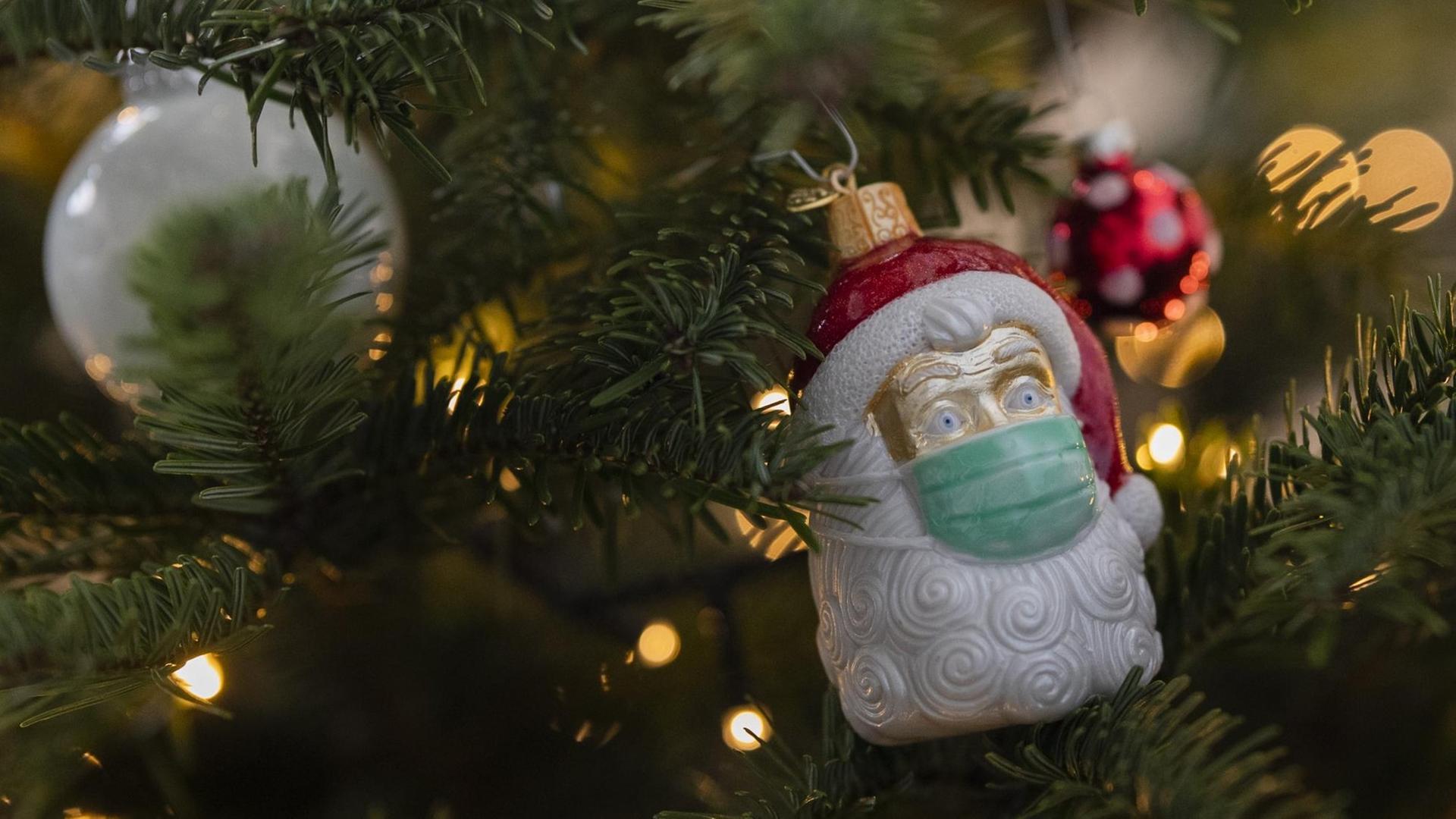 An einem Weihnachtsbaum hängt Weihnachtsschmuck in Form eines Weihnachtsmannes, der eine Maske trägt.