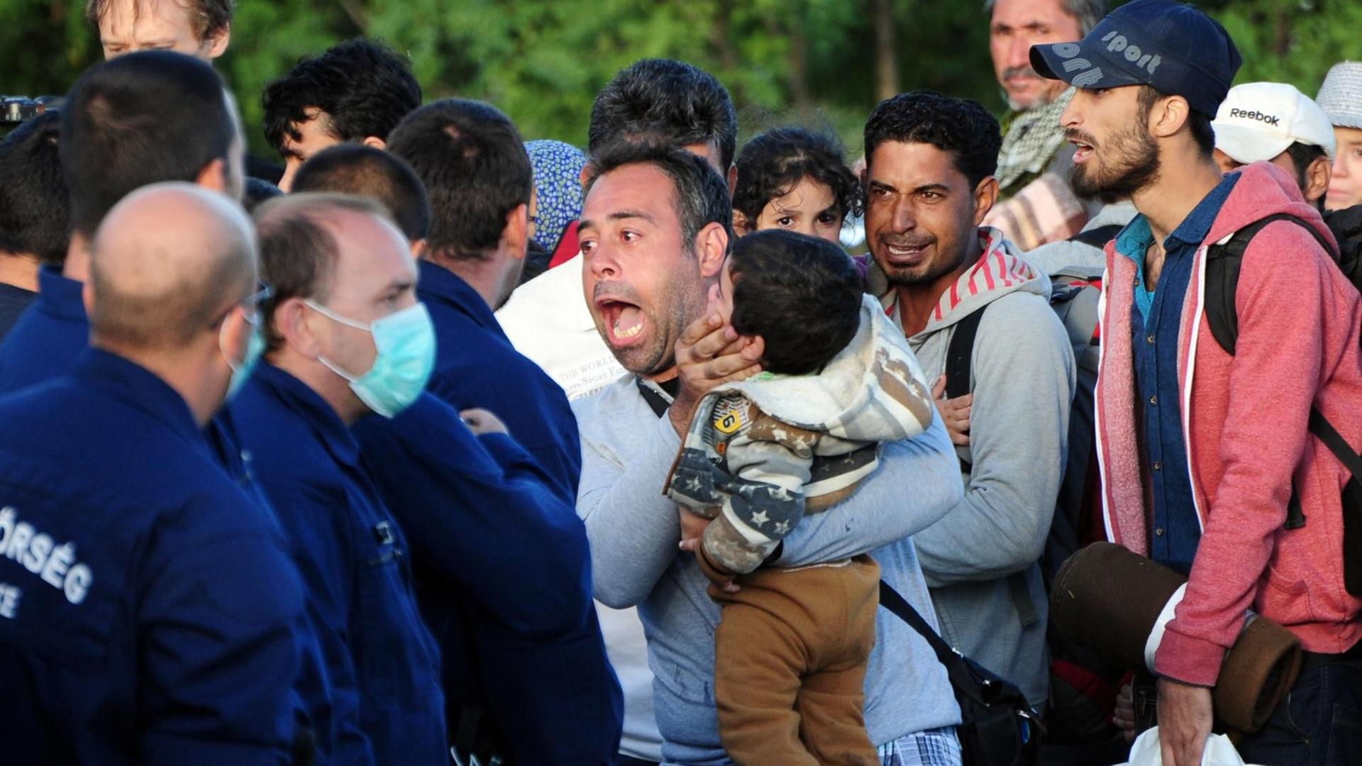 Polizisten in Uniform und zum Teil mit Mundschutz stehen aufgebrachten Flüchtlingen gegenüber. Ein Mann schreit die Polizisten an, hält ein Kind auf dem Arm.