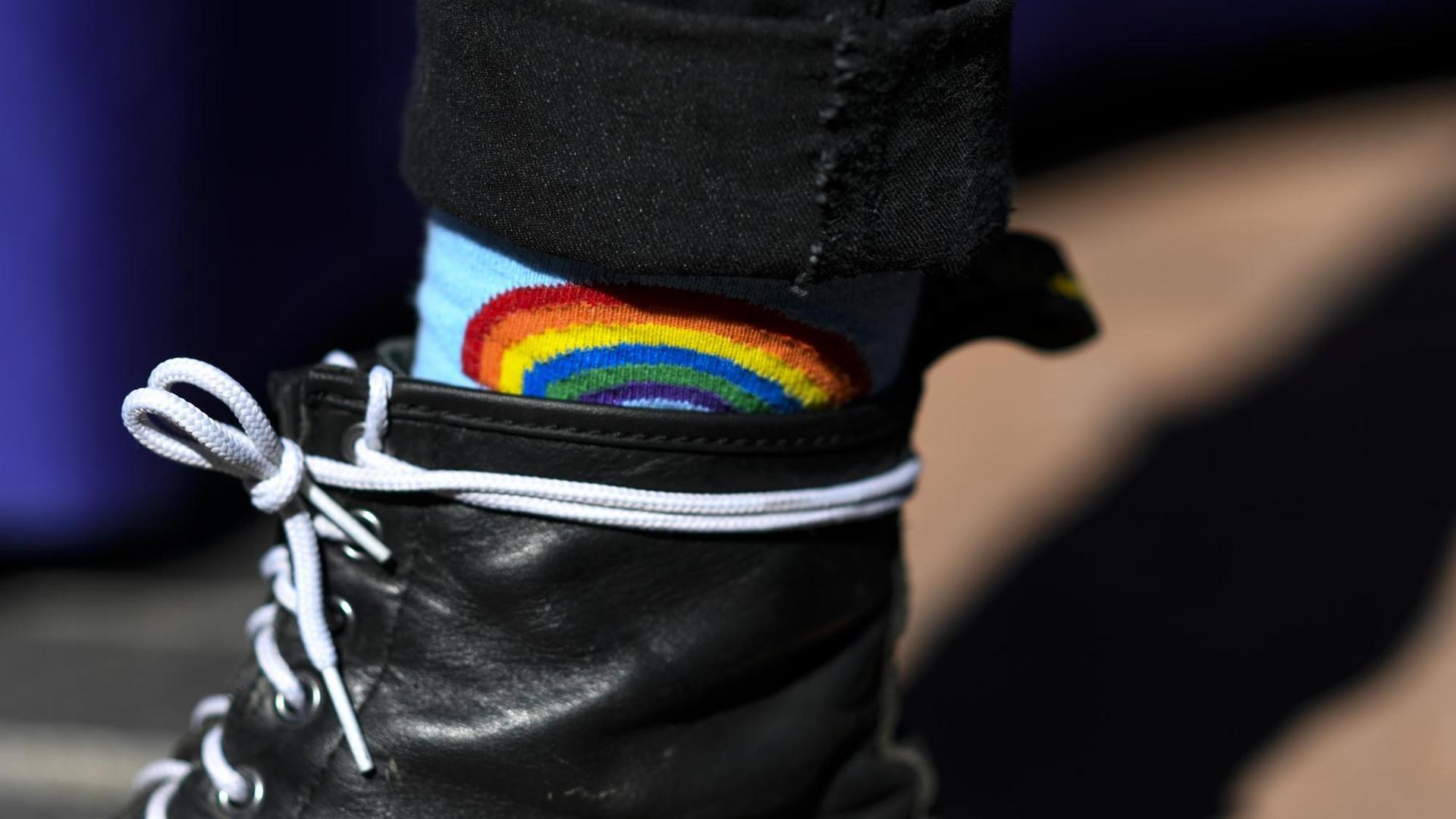 Detailaufnahme einer Regenbogen-Socke, die zwischen schwarzen Stiefeln und Hosen herausschaut.