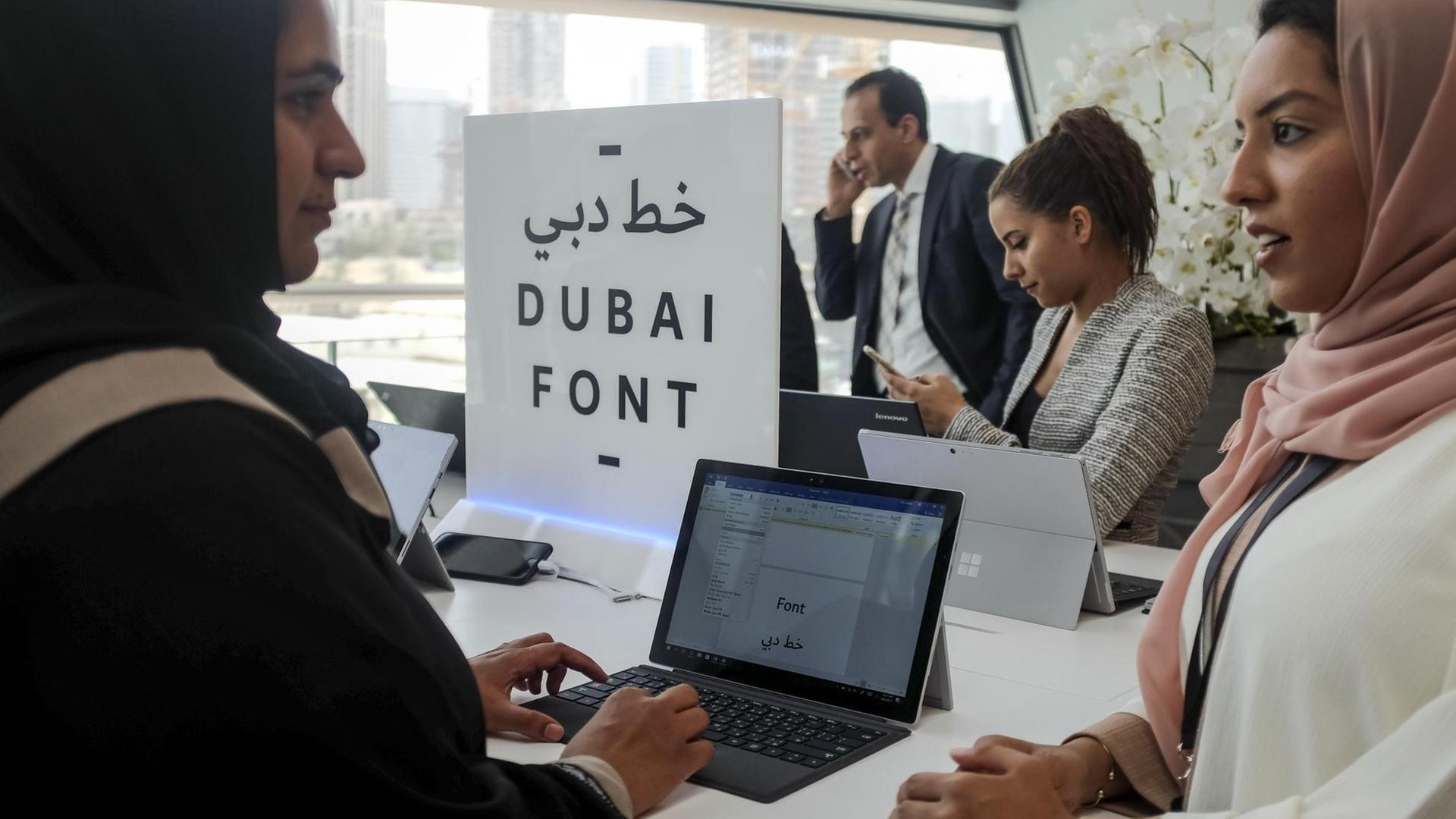 Sie sehen drei Frauenan Laptops, in der Mitte ein Schild mit der Aufschrift "DUBAI FONT". Im Hintergrund telefoniert ein Mann.