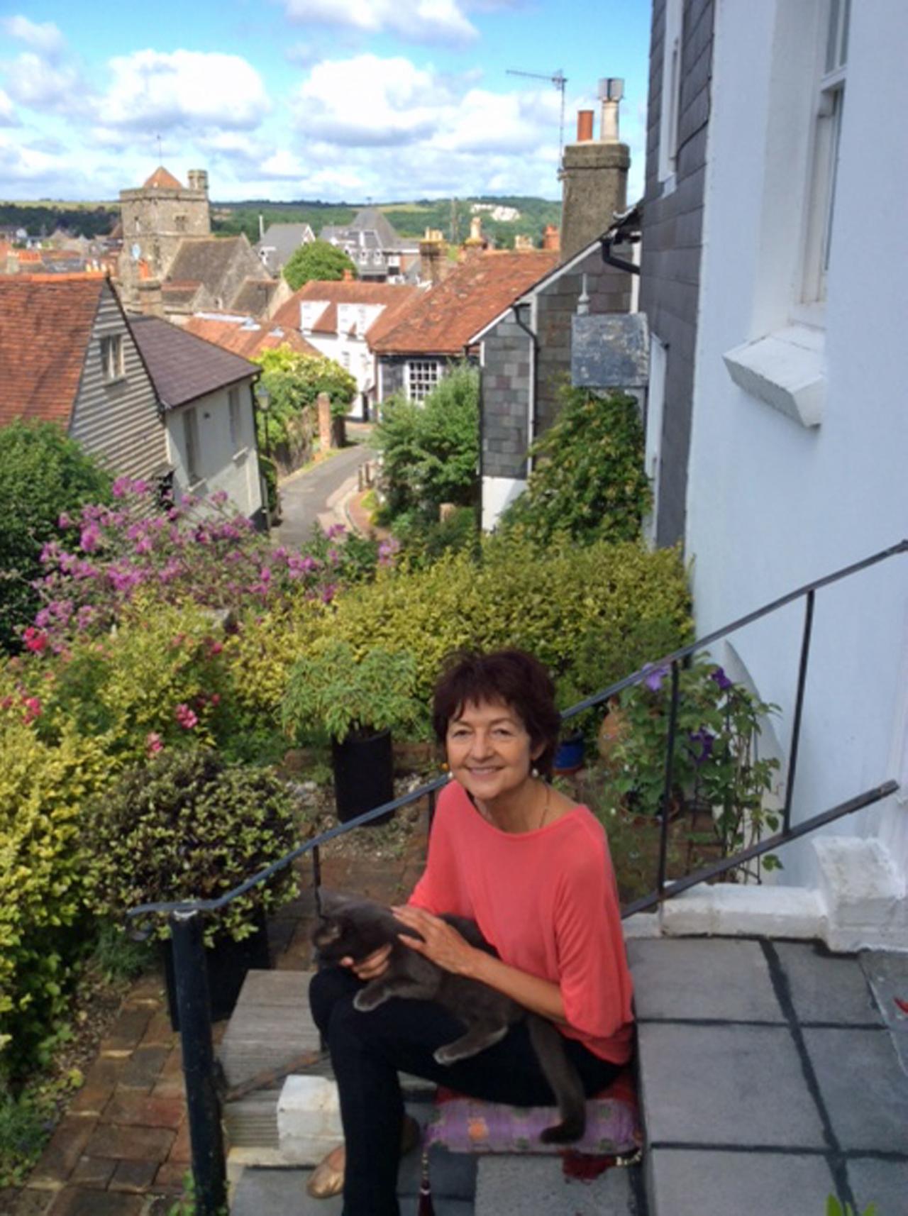 Farbfoto einer Frau in mittleren Jahren, die eine Katze auf dem Schoß hat. Sie sitzt auf einer Treppe, im Hintergrund ist eine Stadtlandschaft zu sehen. Die Frau lächelt.