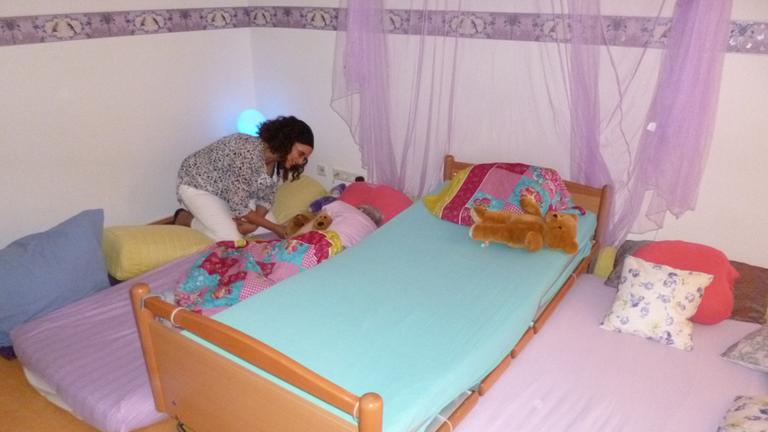 Pflegerin Nora Moatassimi mit Heimbewohnerin Anni. Zimmer mit Bett und zwei Matratzen auf dem Boden, in lila gehalten, auf einer Matratze liegt eine alte Frau, eine jüngere Frau beugt sich über sie.