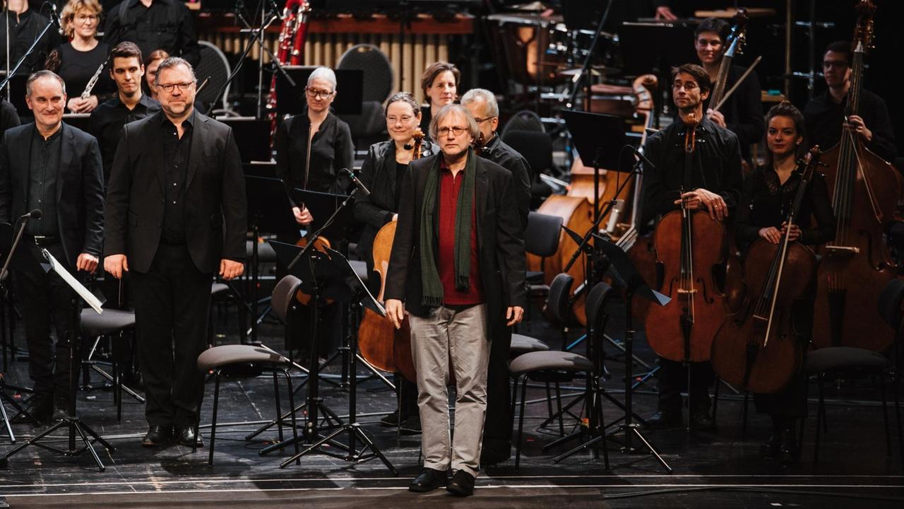 Der Komponist steht auf der Bühne, umringt von Orchester und Sängern