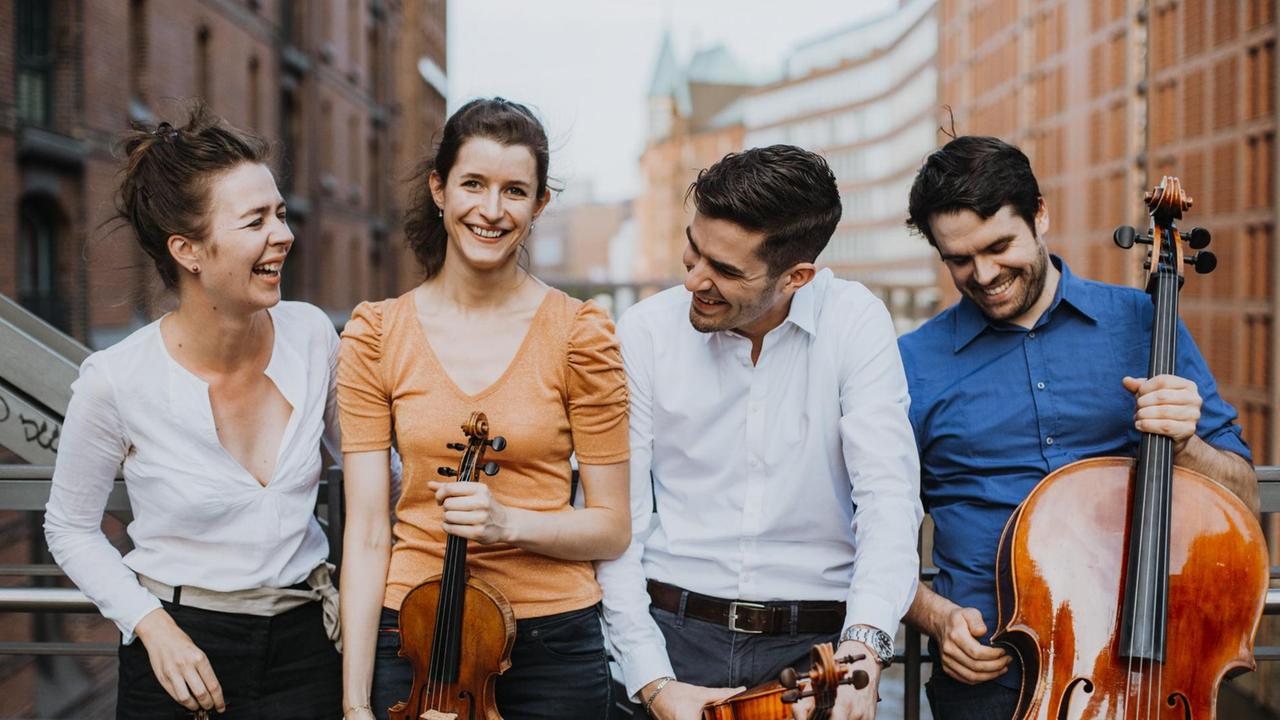 Die vier Musiker, lachend und scherzend, in Alltagskleidung mit ihren Instrumenten auf einer Brücke zwischen hohen Häusern.