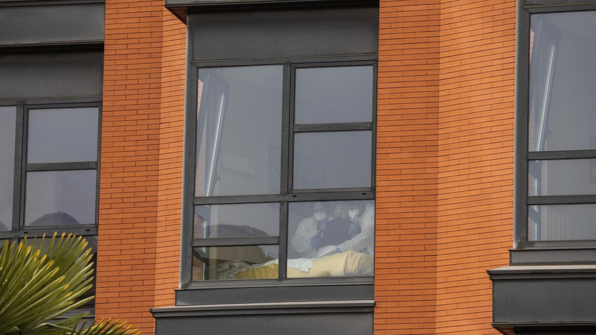 Man sieht die beiden Pflegekräfte und die Seniorin undeutlich durch ein Fenster an der Fassade. Daneben zwei weitere Fenster.
