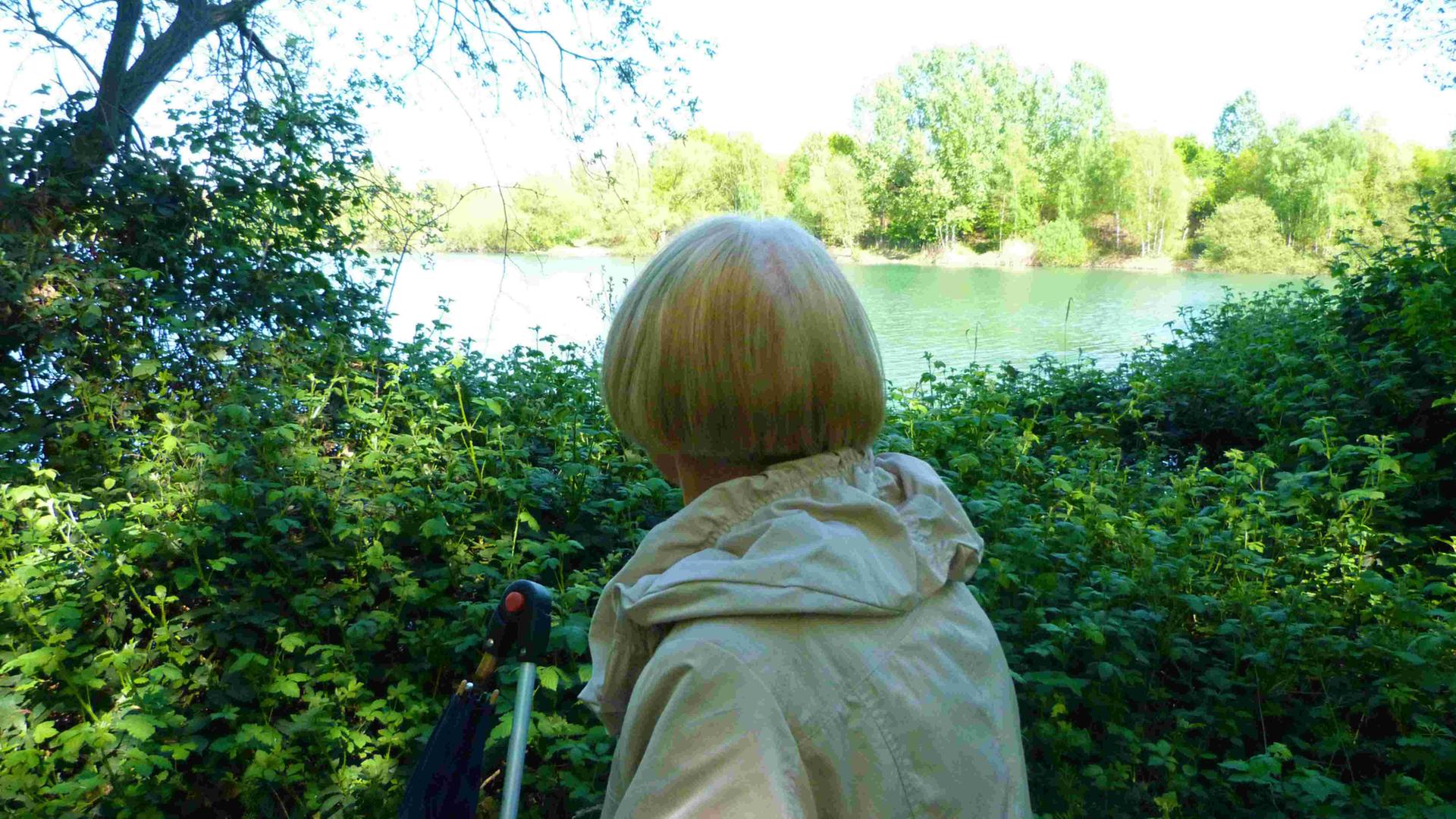 Margot mit dem Rücken zur Kamera. Sie hat halblange blonde Haare, trägt einen grauen Anorak und schaut auf einen See. Margot mit dem Rücken zur Kamera. Sie hat halblange blonde Haare, trägt einen grauen Anorak und schaut auf einen See.