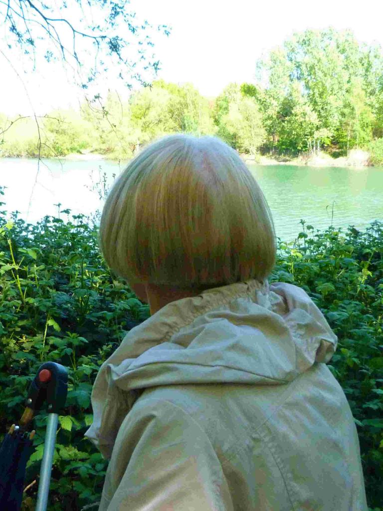 Margot mit dem Rücken zur Kamera. Sie hat halblange blonde Haare, trägt einen grauen Anorak und schaut auf einen See.