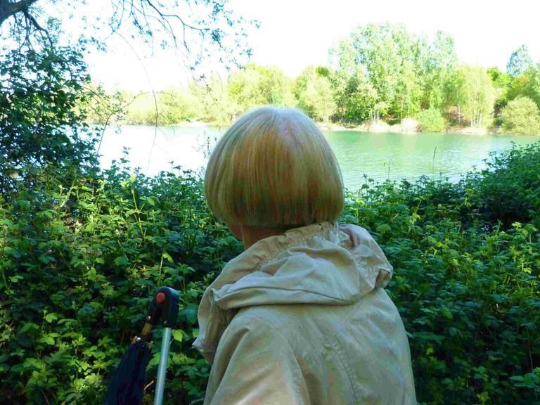 Margot mit dem Rücken zur Kamera. Sie hat halblange blonde Haare, trägt einen grauen Anorak und schaut auf einen See.