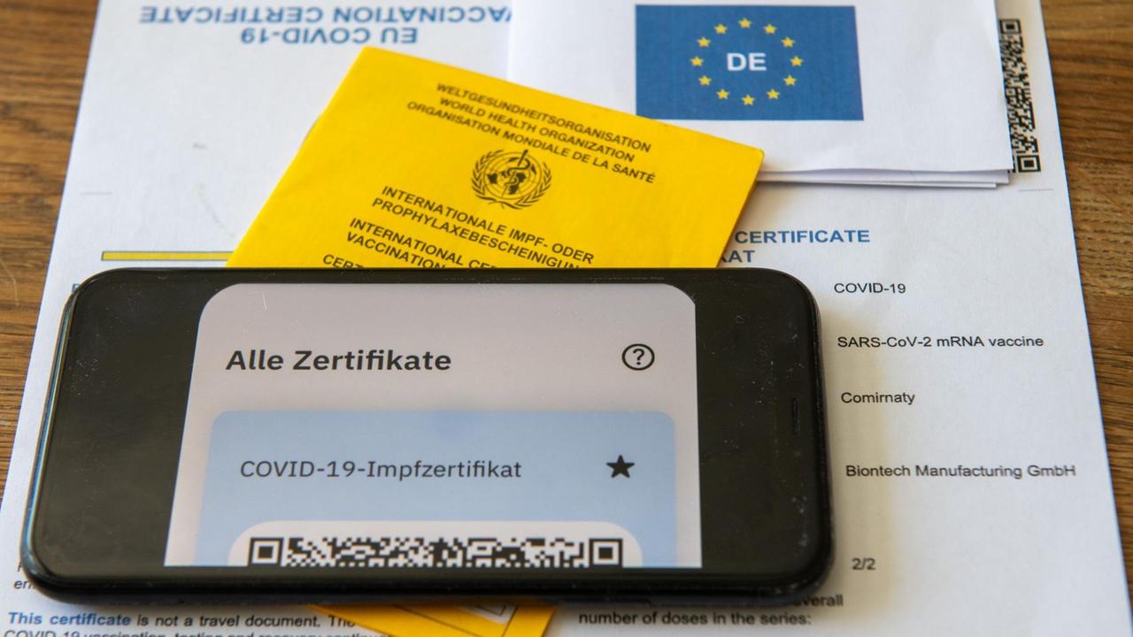 Ein Impfpass und ein Smartphone, auf dem die App CovPass läuft, liegen auf einem Impfzertifikat, das von einer Apotheke ausgestellt wurde. Der digitale Nachweis ist eine freiwillige Ergänzung des weiter gültigen gelben Impfheftes aus Papier. Deutschland setzt damit ein Vorhaben der Europäischen Union um.