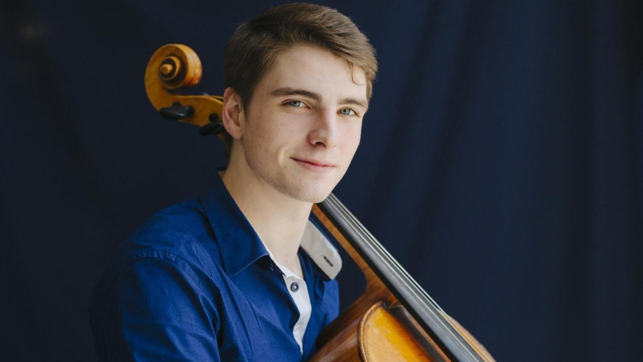 Ein junger Mann mit kurzen, hellbraunen Haaren schaut auf einer Porträtaufnahme die Kamera, an seiner linken Schulter lehnt der Hals eines Cellos.