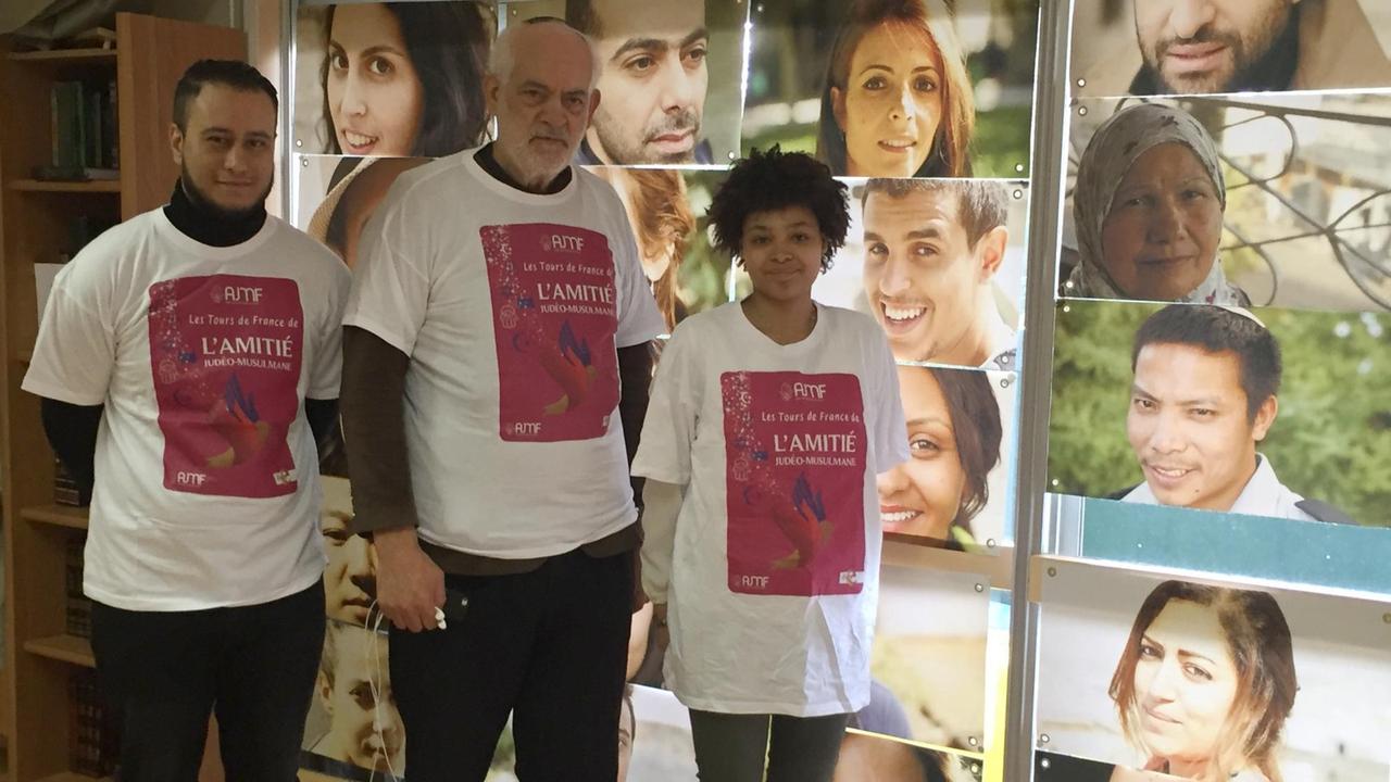 Mohamed, Rabbi Serfaty und Isadora (v.l.n.r) stehen in T-Shirts des Freundschaftsvereins vor einer Fotowand mit Porträts von Männern und Frauen.