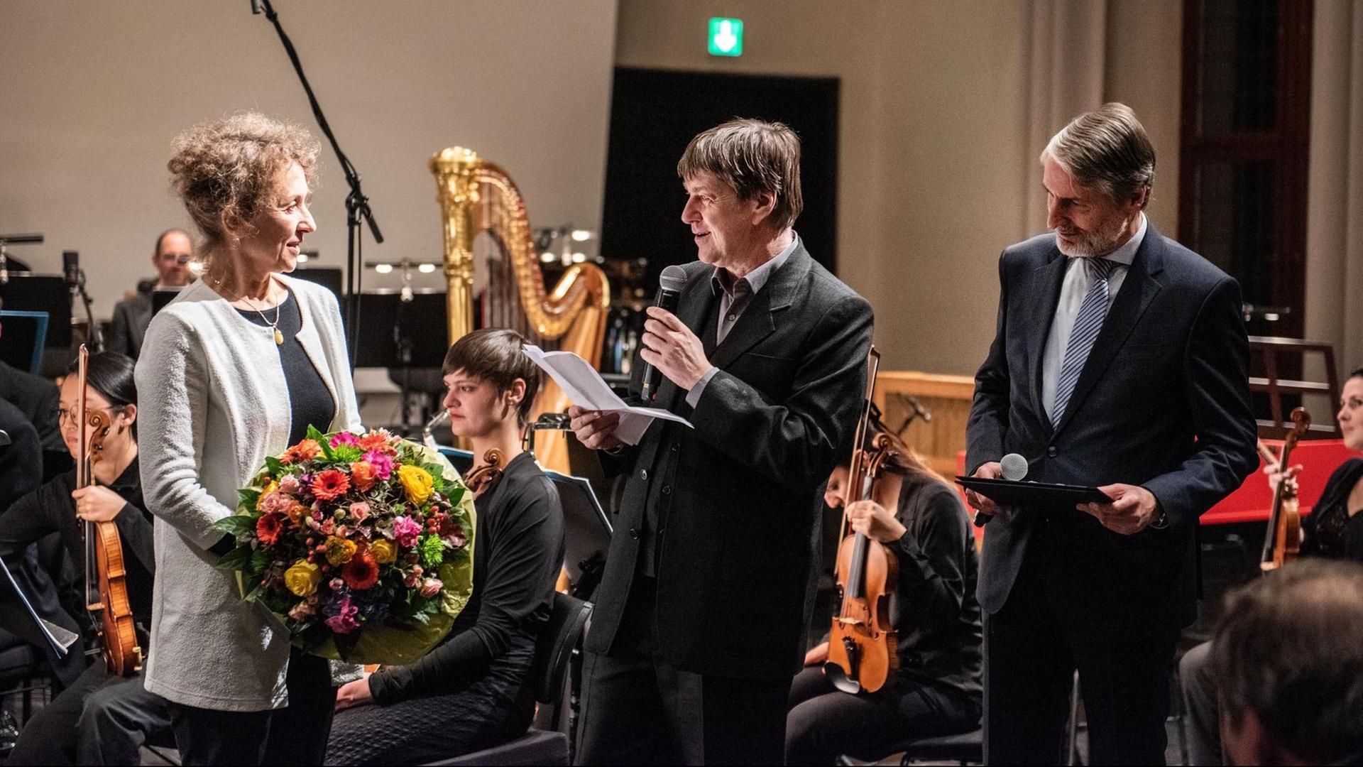 Links hält eine Frau einen Blumenstrauß in der Hand, rechts von ihr stehen zwei Herren im Anzug mit Mikrofon in der Hand, einer davon hält eine Ansprache. Im Hintergrund ist ein Orchester im Ausschnitt zu sehen.