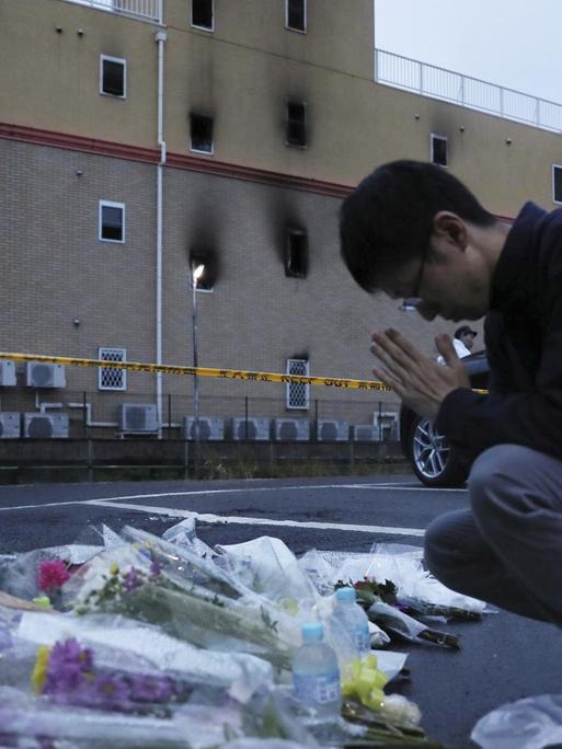 Ein Mann mit gefalteten Händen kniet vor dem Kyoto-Animation-Studio. Vor ihm liegen Blumen. Das Gebäude im Hintergrund hat Rußspuren.