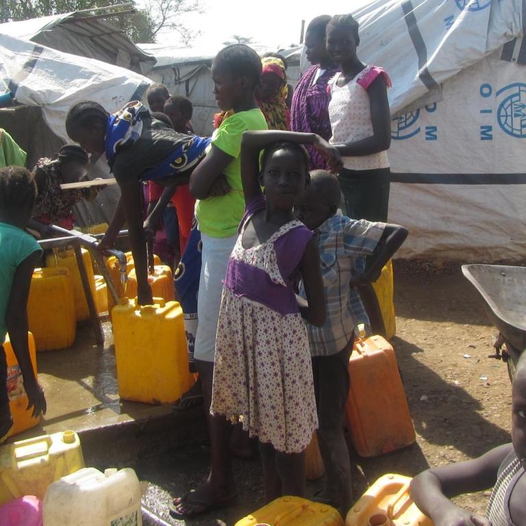 Frauen und Kinder stehen mit Eimern an mehreren Wasserhähnen im Flüchtlingslager.
