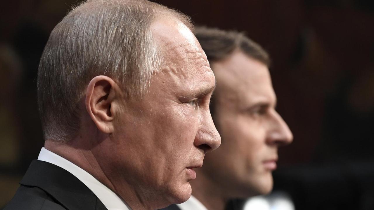 Nahaufnahme von Putins Kopf von der Seite, dahinter unscharf der Kopf Macrons vor einem dunklen Hintergrund.
