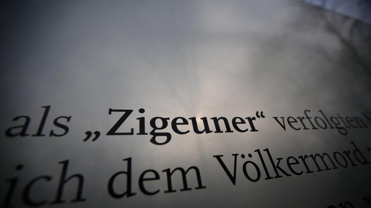 Zigeuner steht auf einer Hinweistafel am Mahnmal, während durch de Glassscheibe die Bäume zu sehen sind.
