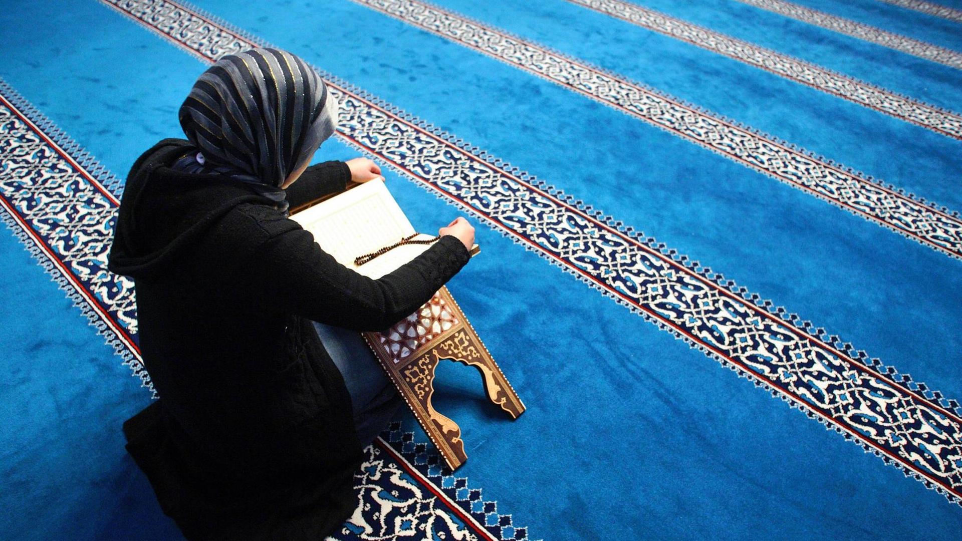 Eine Frau liesst in der Sehitlik Moschee am Columbiadamm in Berlin-Neukölln im Koran. Sie ist schwarz gekleidet, der Teppich in der Moschee ist überwiegend blau.