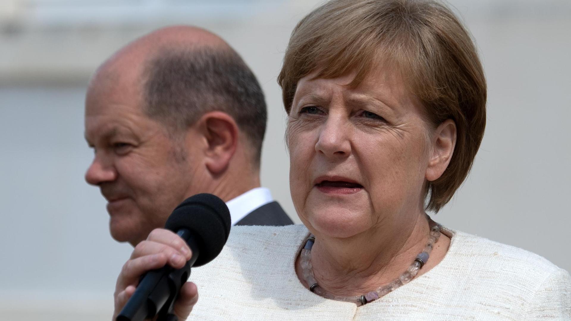 Bundeskanzlerin Angela Merkel (CDU) mit Mikrophon in der Hand, im Hintergrund Olaf Scholz (SPD) und das Gästehaus der Bundesregierung in Meseberg