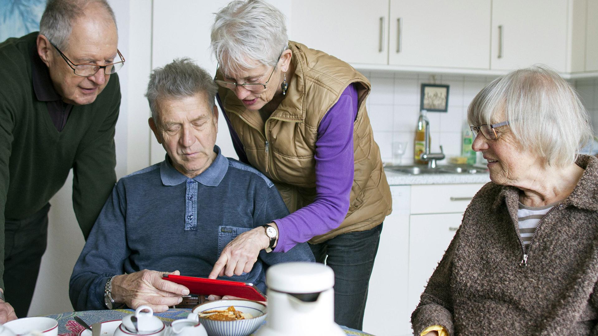Vier ältere Menschen beim gemeinsamen Kaffeetrinken in der Küche schauen auf ein rotes Tablet