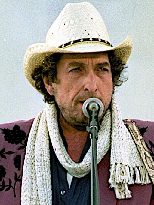 Bob Dylan vor einem Mikrofon. Er rtägt eine rotbraune Jacke, einen weißen Schal und einen weißen Hut.