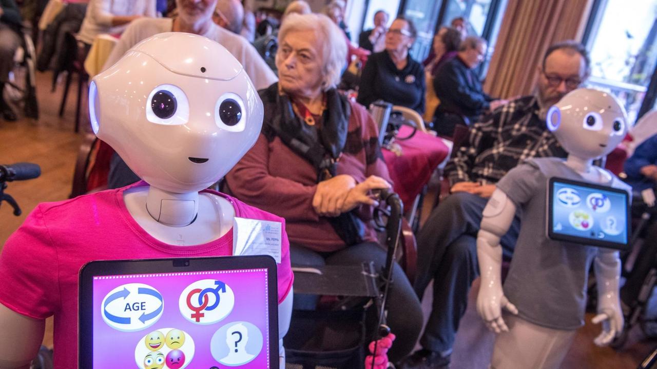 Zwei menschenähnliche Roboter stehen mit bunten T-Shirts zwischen älteren Menschen im Altenpflegeheim