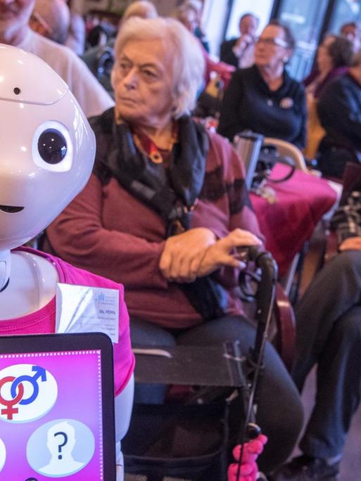 Zwei menschenähnliche Roboter stehen mit bunten T-Shirts zwischen älteren Menschen im Altenpflegeheim